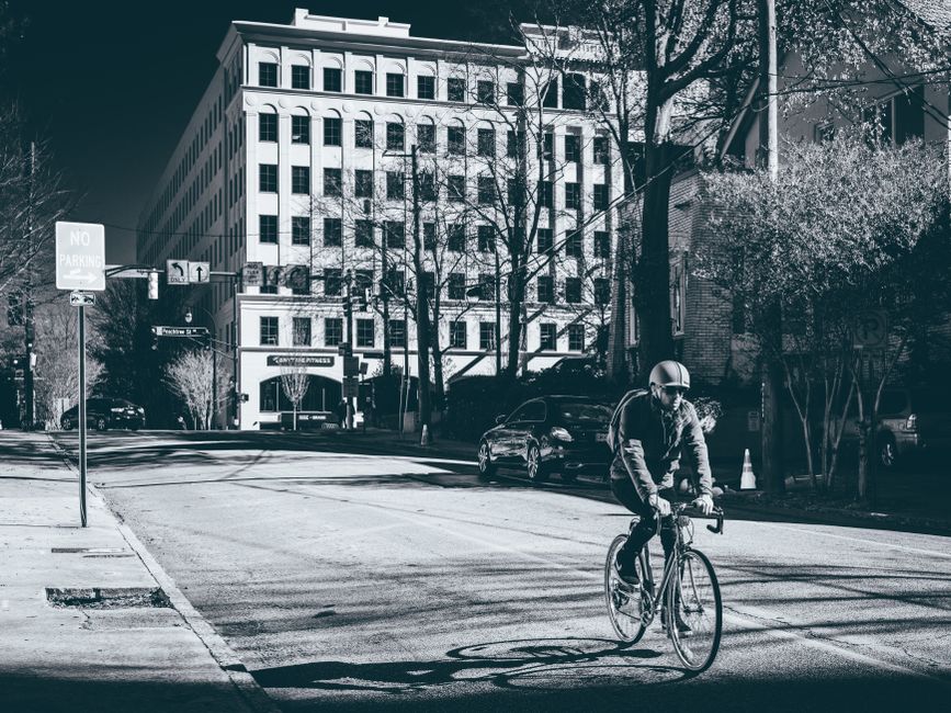 A cyclist wearing a helmet rides a bicycle along a city street near a large building.